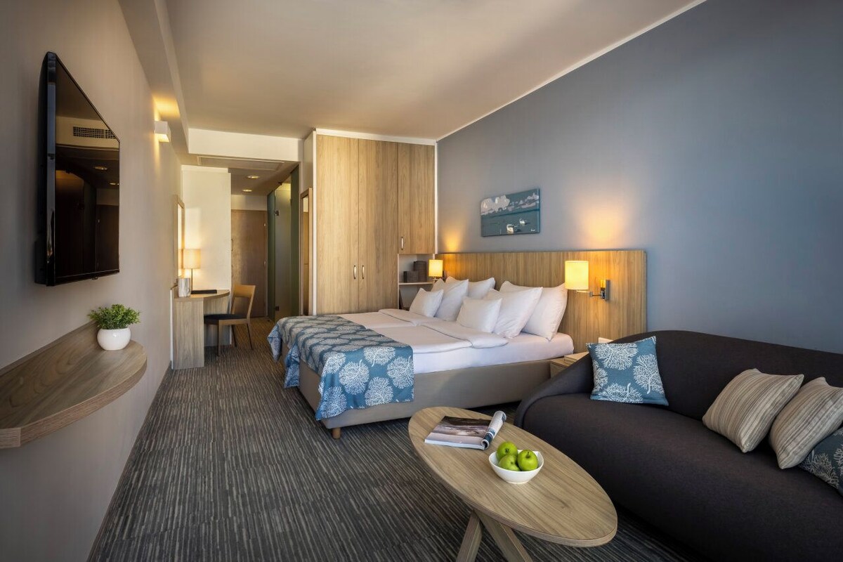 Habitación de hotel con cama, sofá, una mesita con manzanas, televisión montada en la pared y un mueble de madera.
