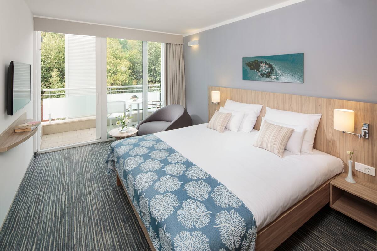 A hotel room with a large bed, a balcony with a view of greenery, a chair, and a small table.
