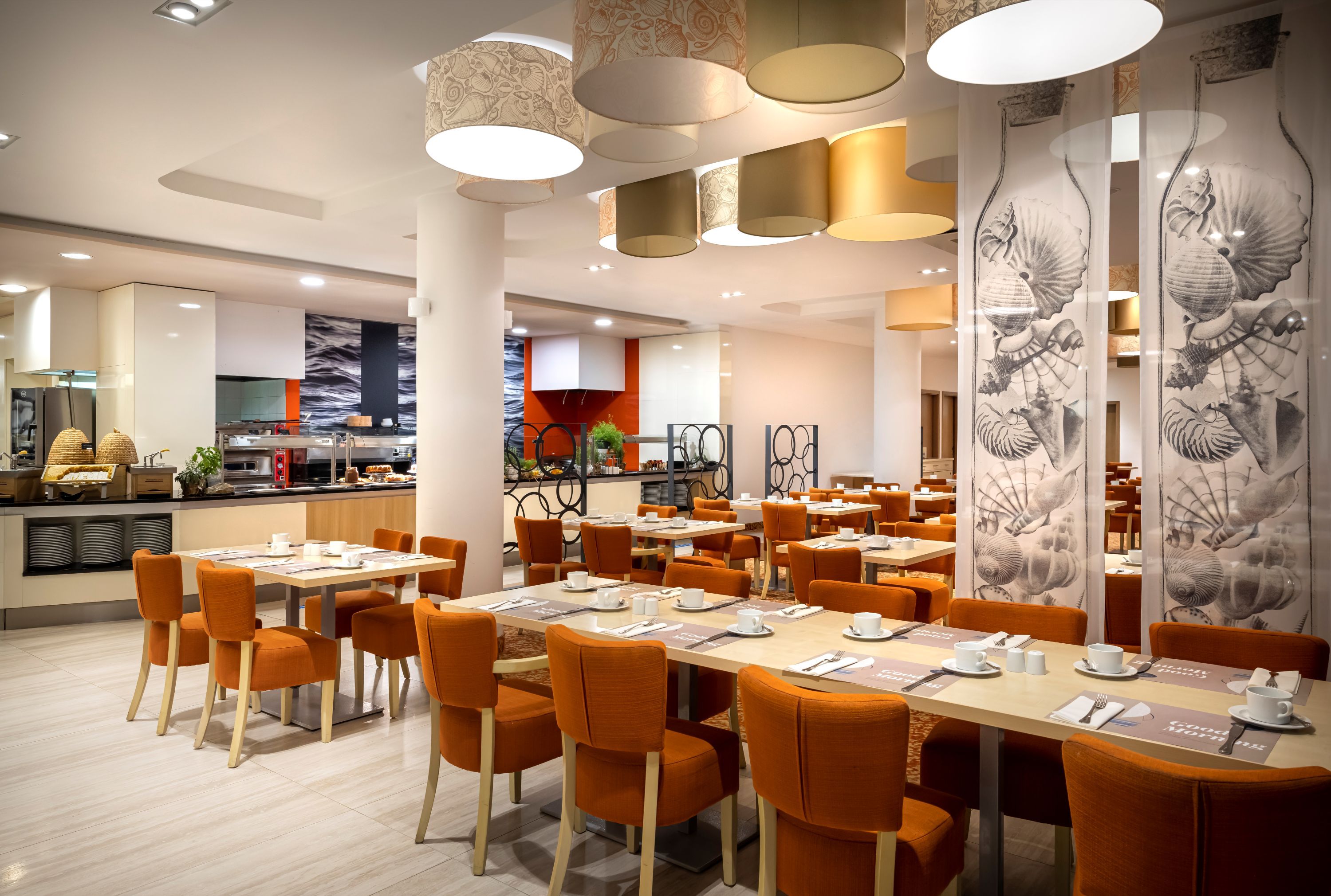 A modern dining area with multiple tables and chairs, overhead lighting, and a kitchen in the background.