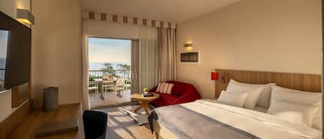 A hotel room with a large bed, a desk with a chair, a TV, and a balcony with outdoor furniture.