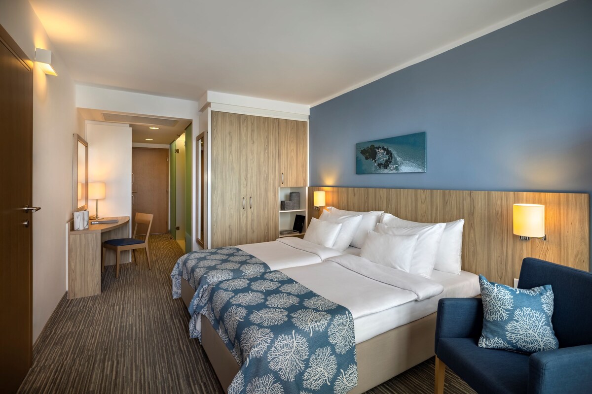 Una habitación de hotel con una cama grande, dos mesitas de noche, un escritorio, una silla y una pared azul con un cuadro.