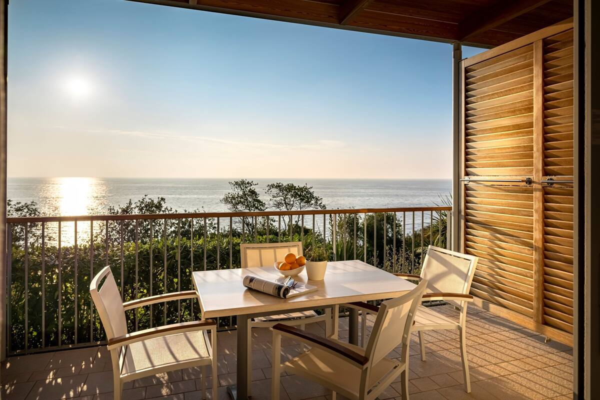 A balcony with a table and chairs, a bowl of fruit, and a view of the sea.