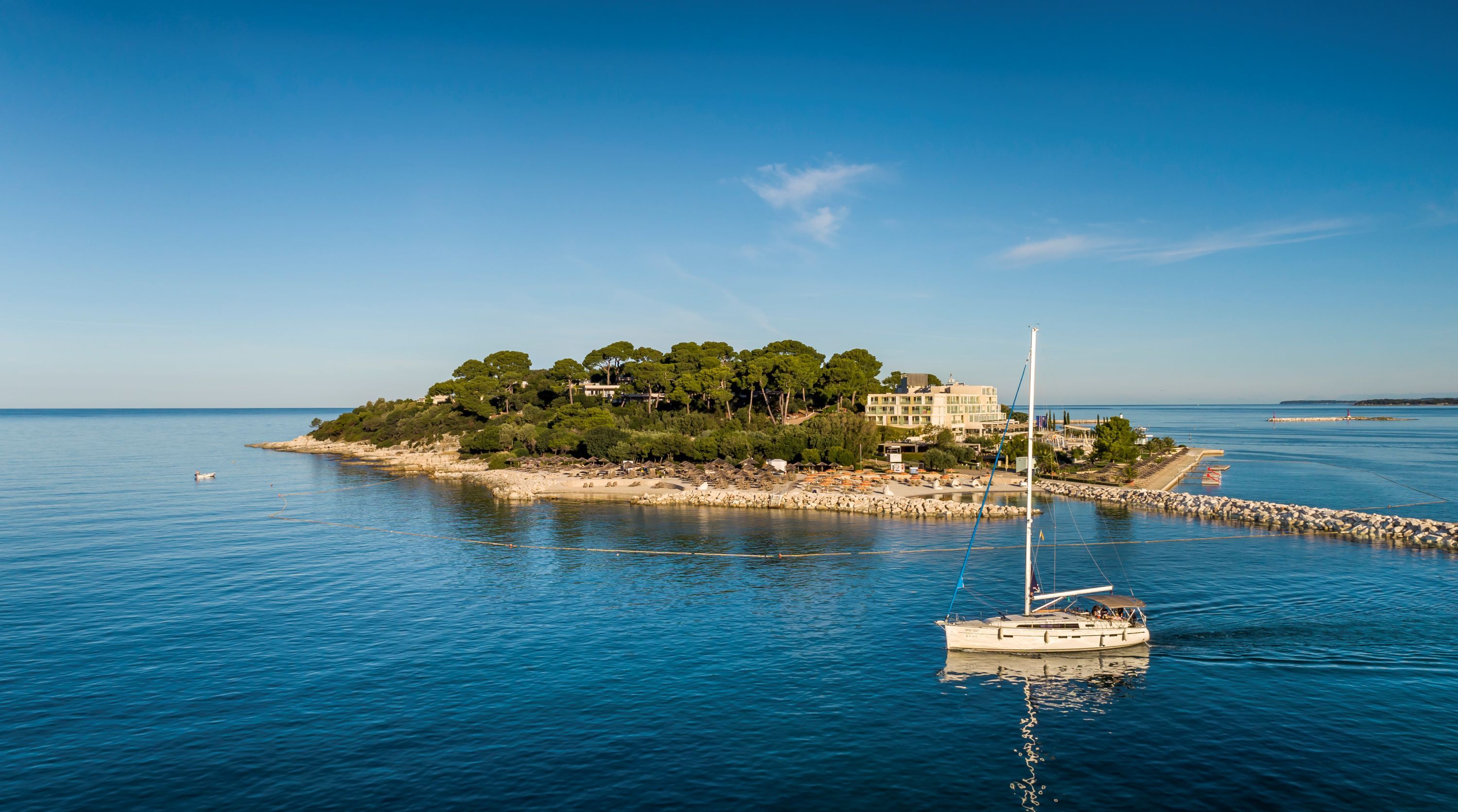 A coastal scene with a small island featuring a building surrounded by trees, a rocky shoreline, and a sailboat in the water.