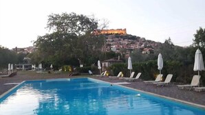 Ein Swimmingpool mit Liegestühlen und Sonnenschirmen, umgeben von Grünflächen und einem Hügel mit Gebäuden im Hintergrund.