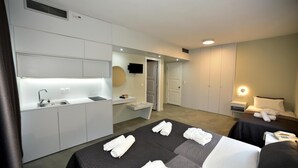 Ein modernes Hotelzimmer mit Küchenzeile, einem Bett mit ordentlich gefalteten Handtüchern, einem Nachttisch mit Lampe und einem großen Spiegel.