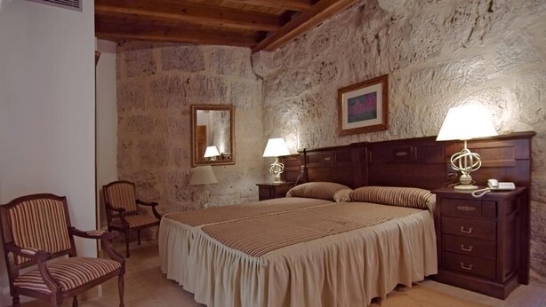 A bedroom with a large bed, two bedside tables with lamps, a wooden chair, and a stone wall.