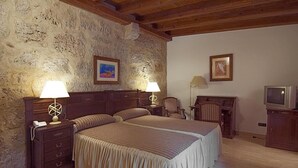 A bedroom with a stone wall, wooden ceiling beams, a double bed, bedside lamps, a chair, a desk, and a television.