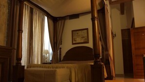 A bedroom with a large bed, wooden posts, curtains, a painting on the wall, and a wooden door.