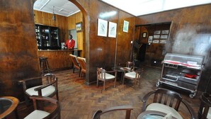Zona de bar con paredes de madera, una barra y taburetes. Se observan cuadros enmarcados en la pared y un carrito con objetos sobre ruedas.