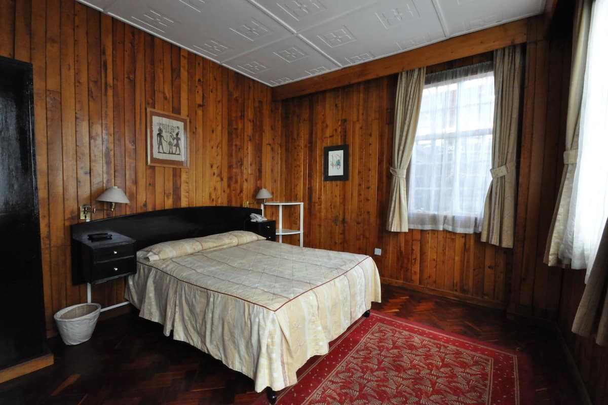 A room with wooden paneling, a bed with a patterned bedspread, a nightstand, a lamp, a framed picture on the wall, and a window with curtains.