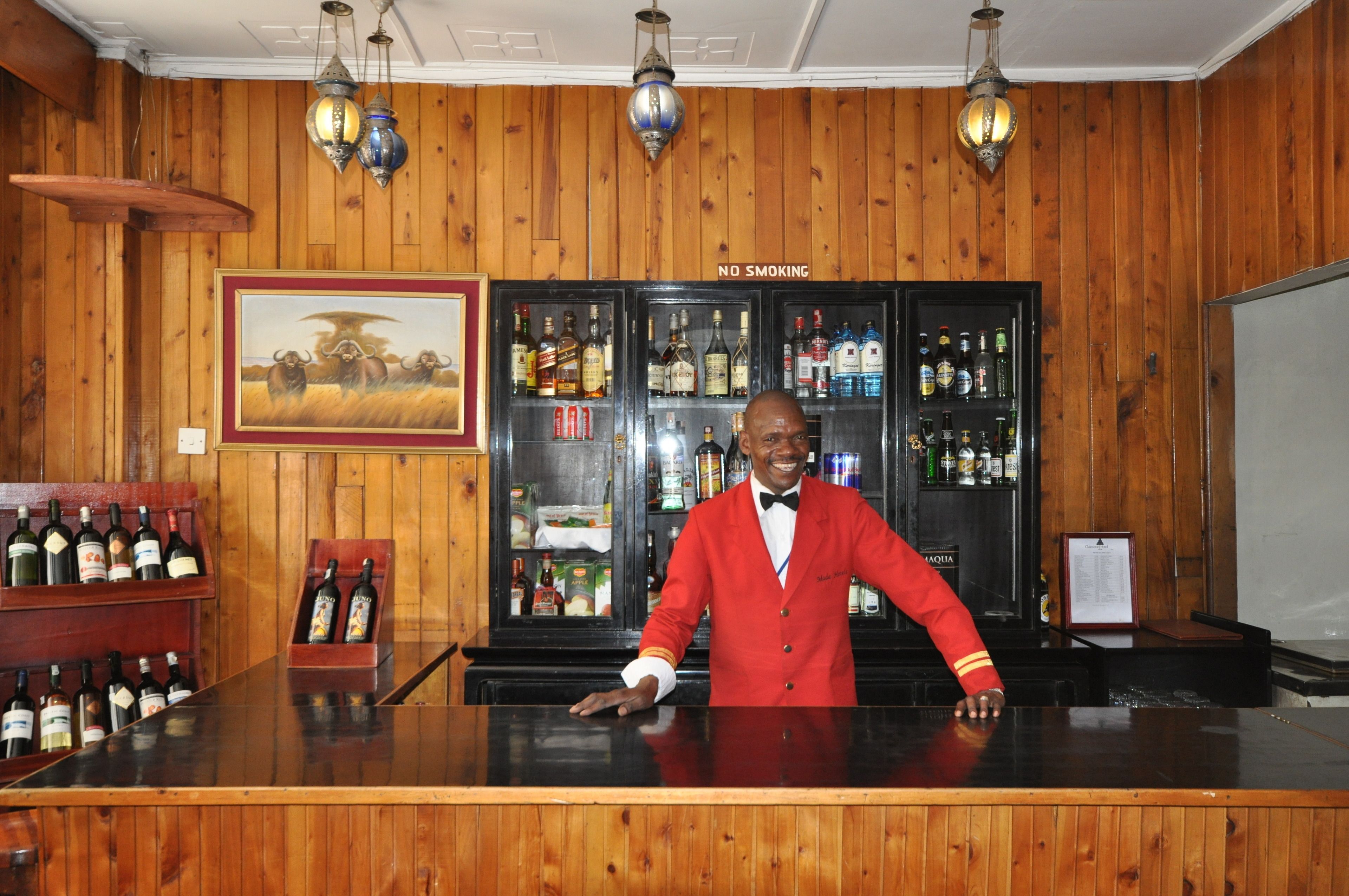 A bar area with wooden paneling, a 'No Smoking' sign, a bartender in a red uniform, and shelves stocked with various liquor bottles.