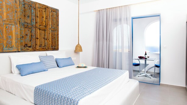 A modern hotel room with a large bed, a wooden wall panel, a hanging lamp, a view of the sea through a glass door, and a small table with a bottle and grapes.