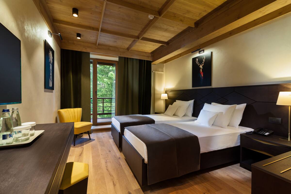 A hotel room with two beds, a desk, a chair, and a view of greenery outside.