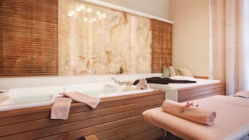 A spa room with a wooden bench, a massage table, and a large mirror with a marble-like backsplash.