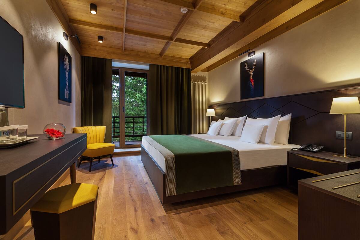 A hotel room with a wooden ceiling, a bed with white linens, a desk with a chair, and a view of greenery outside.