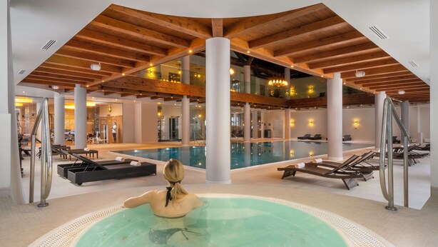 Una amplia piscina interior con un jacuzzi central, rodeada de sillones de descanso y un diseño arquitectónico moderno con vigas de madera y pilares blancos.