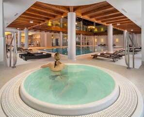 Una amplia piscina interior con un jacuzzi central, rodeada de sillones de descanso y un diseño arquitectónico moderno con vigas de madera y pilares blancos.