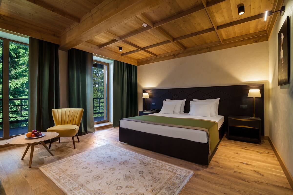 A modern bedroom with a large bed, a sitting area with a chair and table, and a view of greenery through the windows.