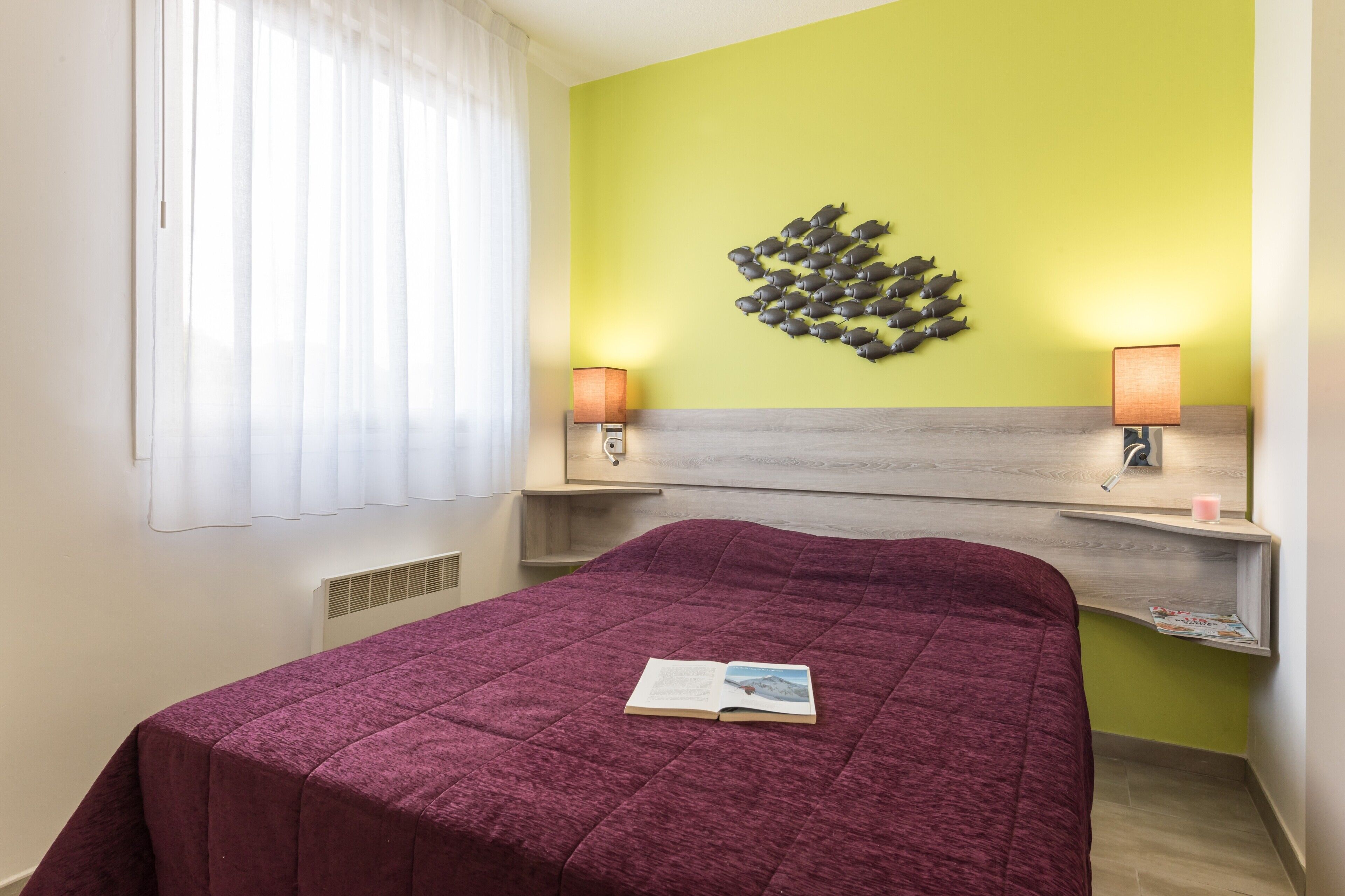 A hotel room with a bed, a headboard, two bedside lamps, a window with sheer curtains, and a wall decoration of fish.