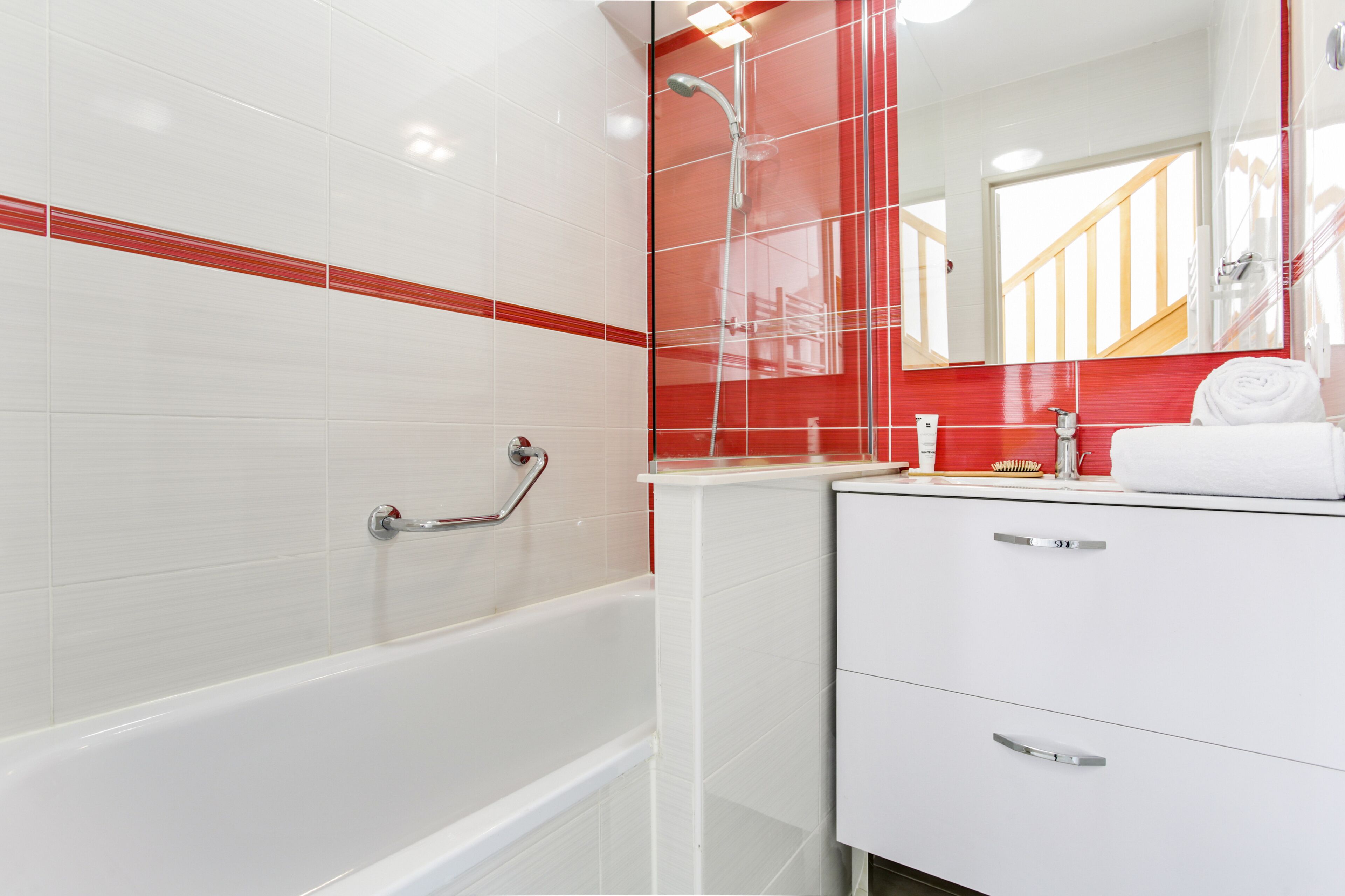 Un baño moderno con una bañera blanca, paredes con azulejos rojos, una ducha con mampara de cristal y un tocador blanco con espejo.