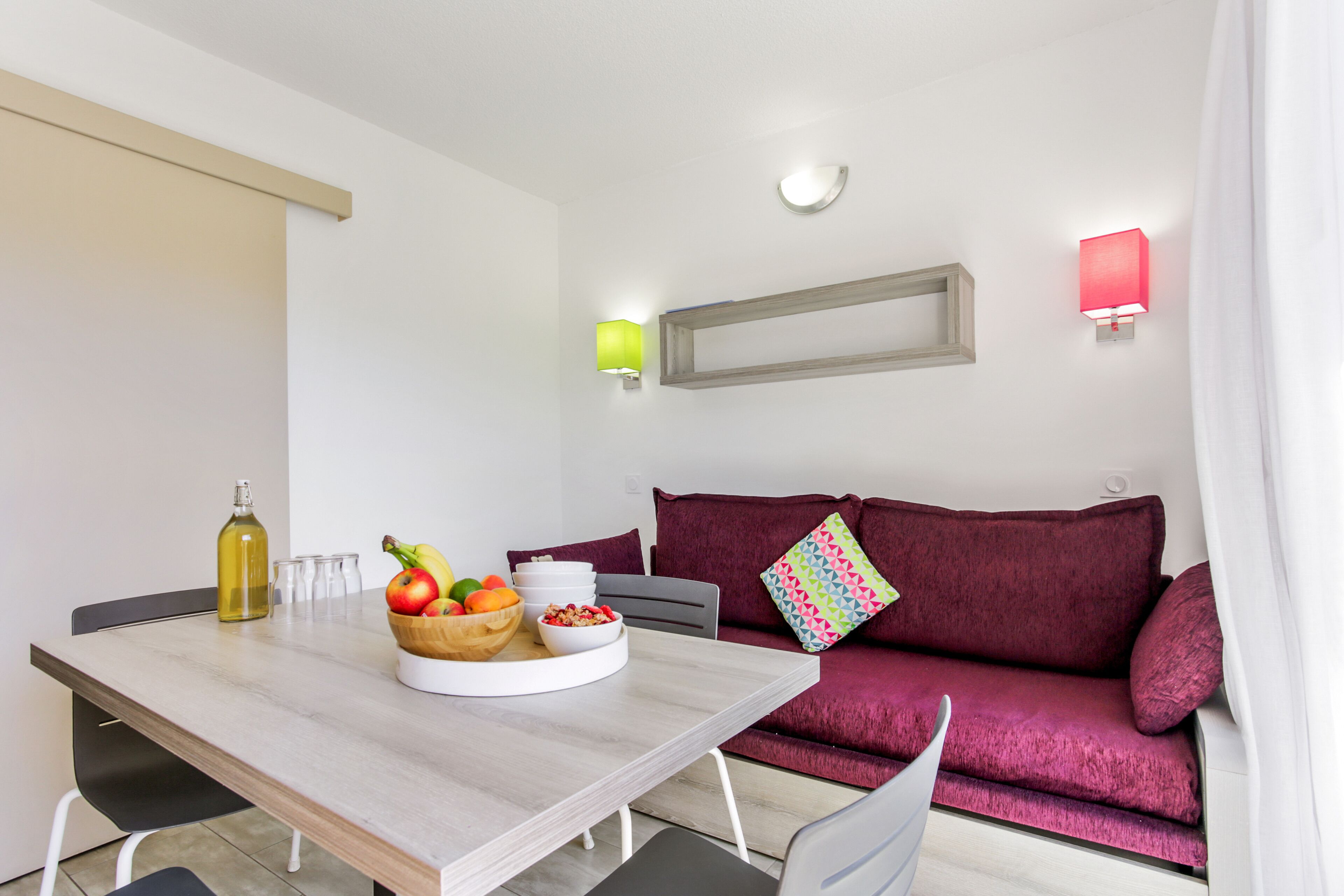 A modern living room with a dining table, a red sofa, and a fruit bowl.