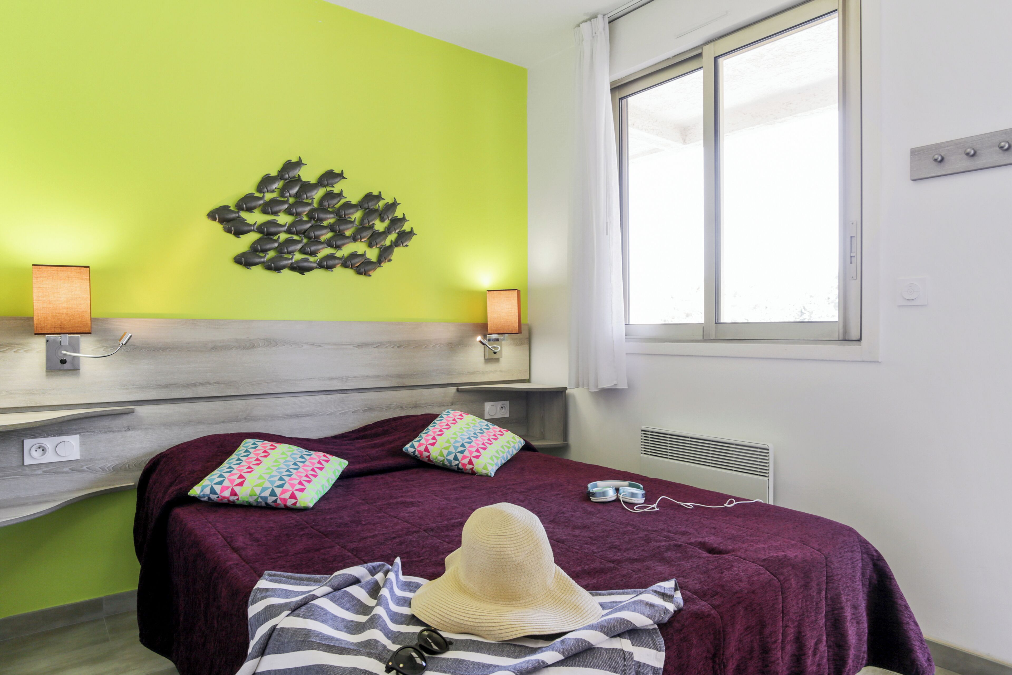 A hotel room with a bed, two wall-mounted lamps, a window with curtains, and a wall art of fish.