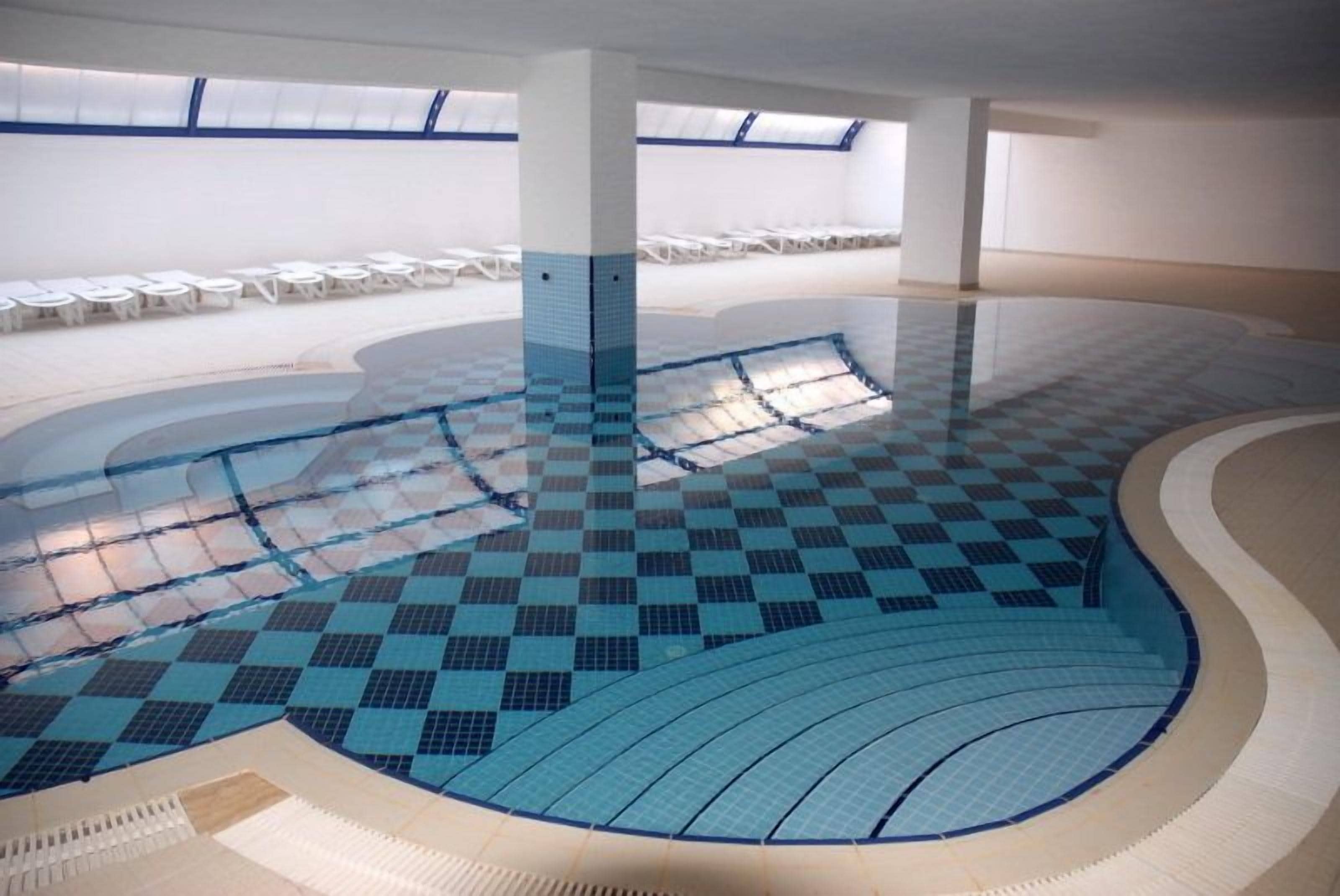 Una piscina interna con pavimento a scacchi, circondata da pareti bianche e un lucernario.