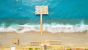 Vista a érea de una playa con un muelle de madera, arena y una estructura de techo plano.