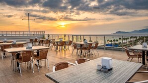 Un área de comedor al aire libre con mesas y sillas, con vista a una playa y al mar.
