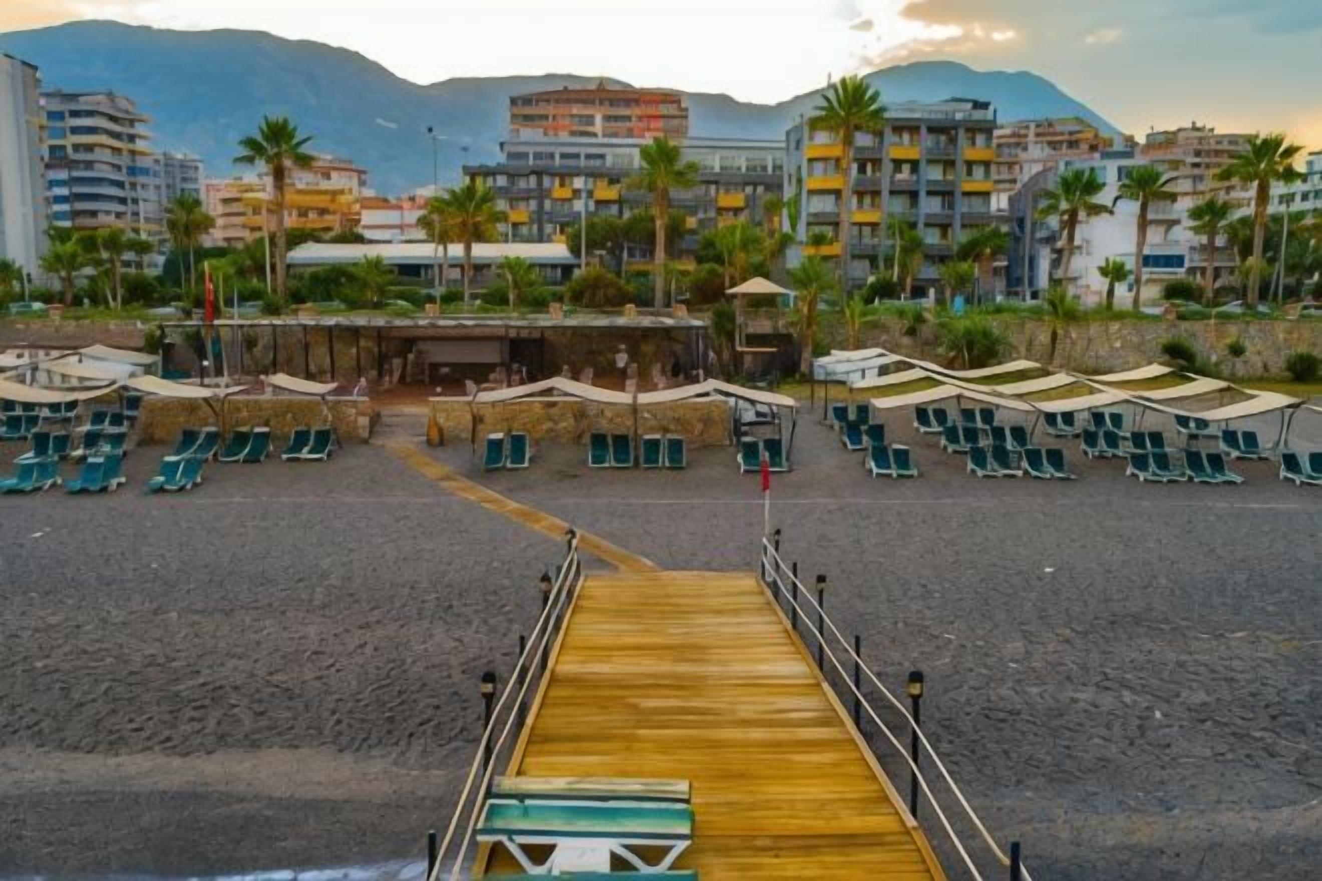 A beach with a wooden pier leading to a row of beach chairs and umbrellas, with buildings and palm trees in the background.