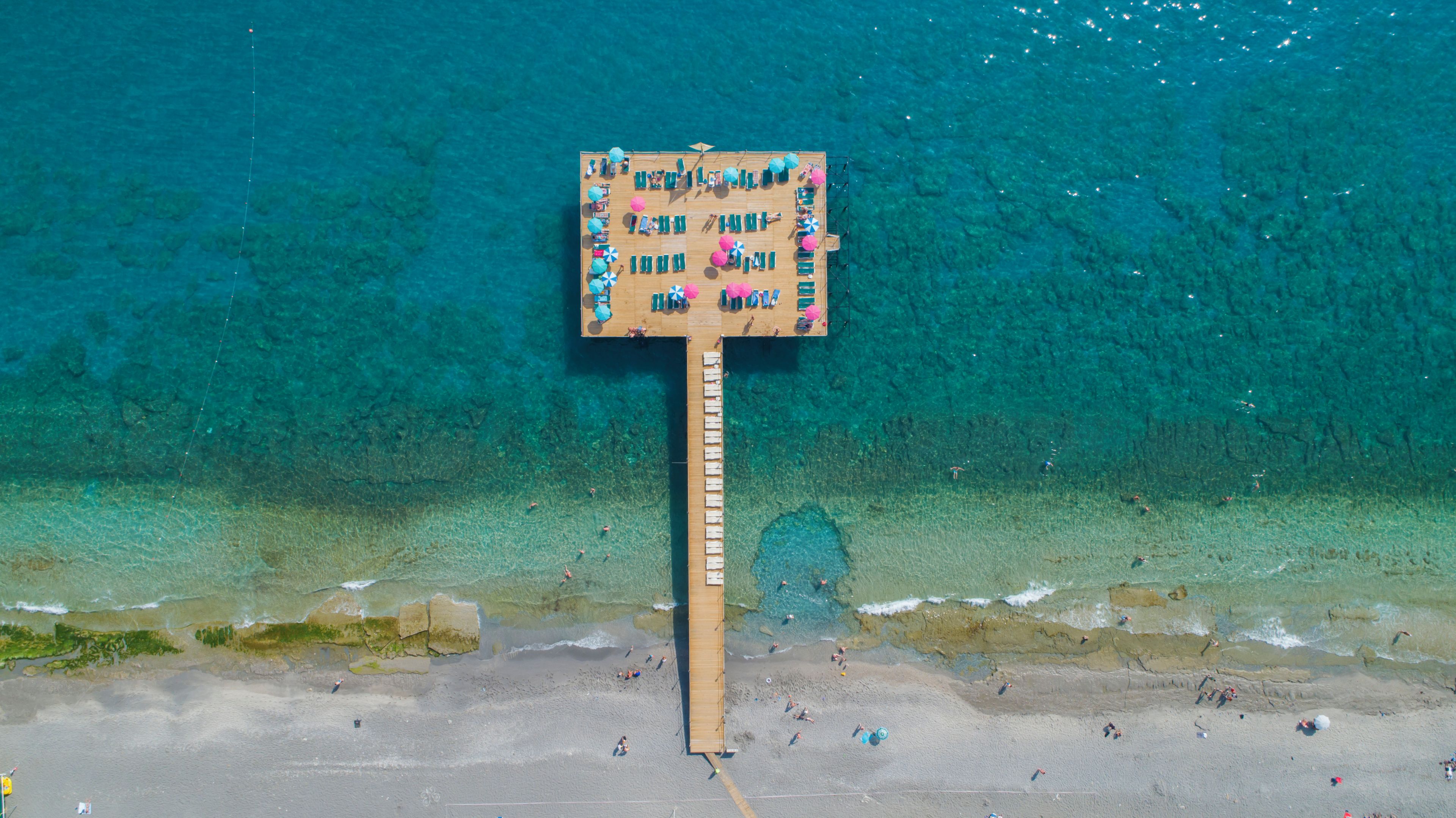 Un pontile in legno con diverse zone relax e ombrelloni, affacciato su una spiaggia con acqua turchese cristallina.