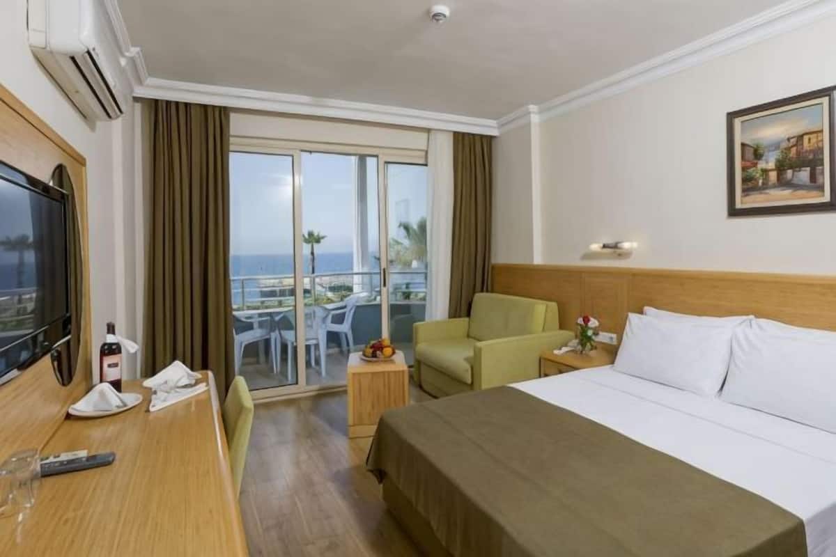 A hotel room with a bed, a desk with a TV, a chair, a small table with a bottle and glasses, and a balcony with a view.