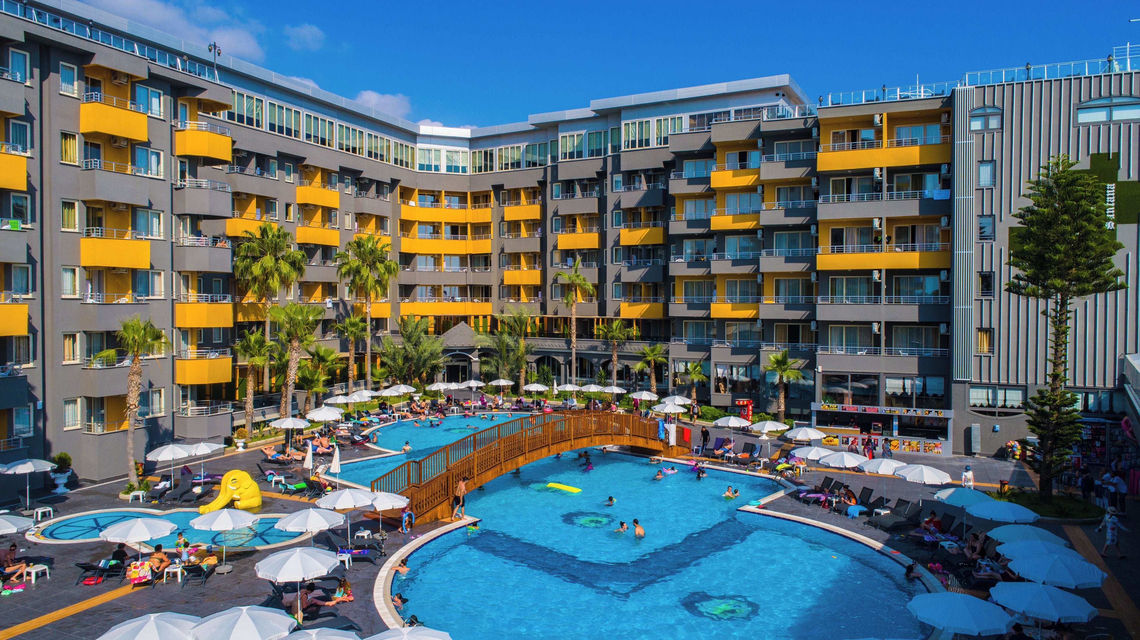 Ett hotell med flera våningar, ett centralt poolområde, flera balkonger och utomhusmöbler.