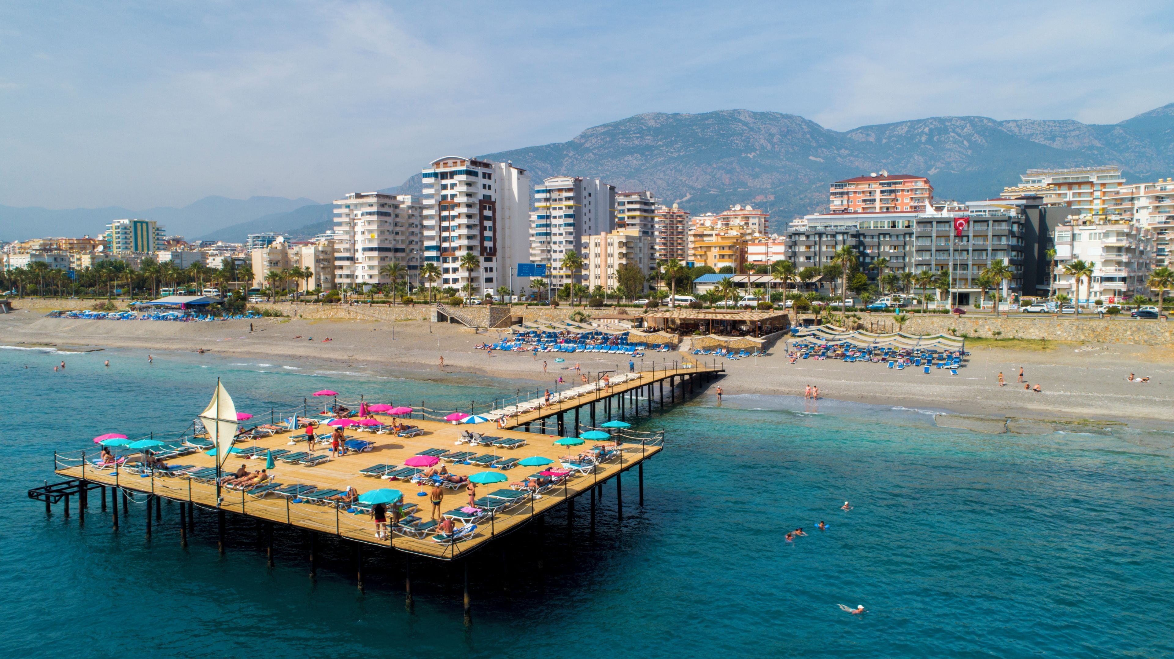 Un paisaje urbano costero con un muelle que se adentra en el mar, bordeado de tumbonas y sombrillas.