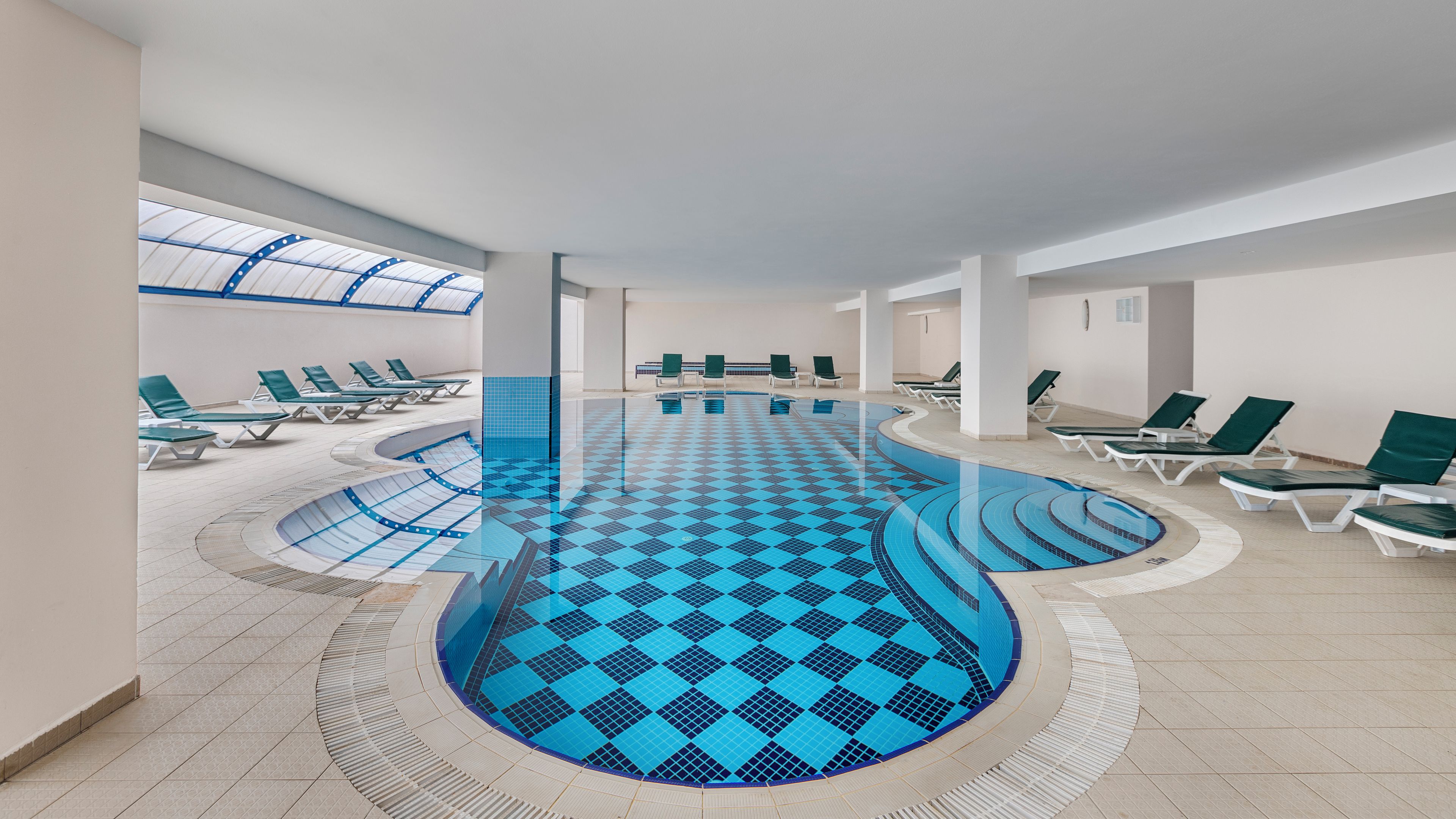 An indoor swimming pool with a blue checkered floor, surrounded by lounge chairs and a skylight above.