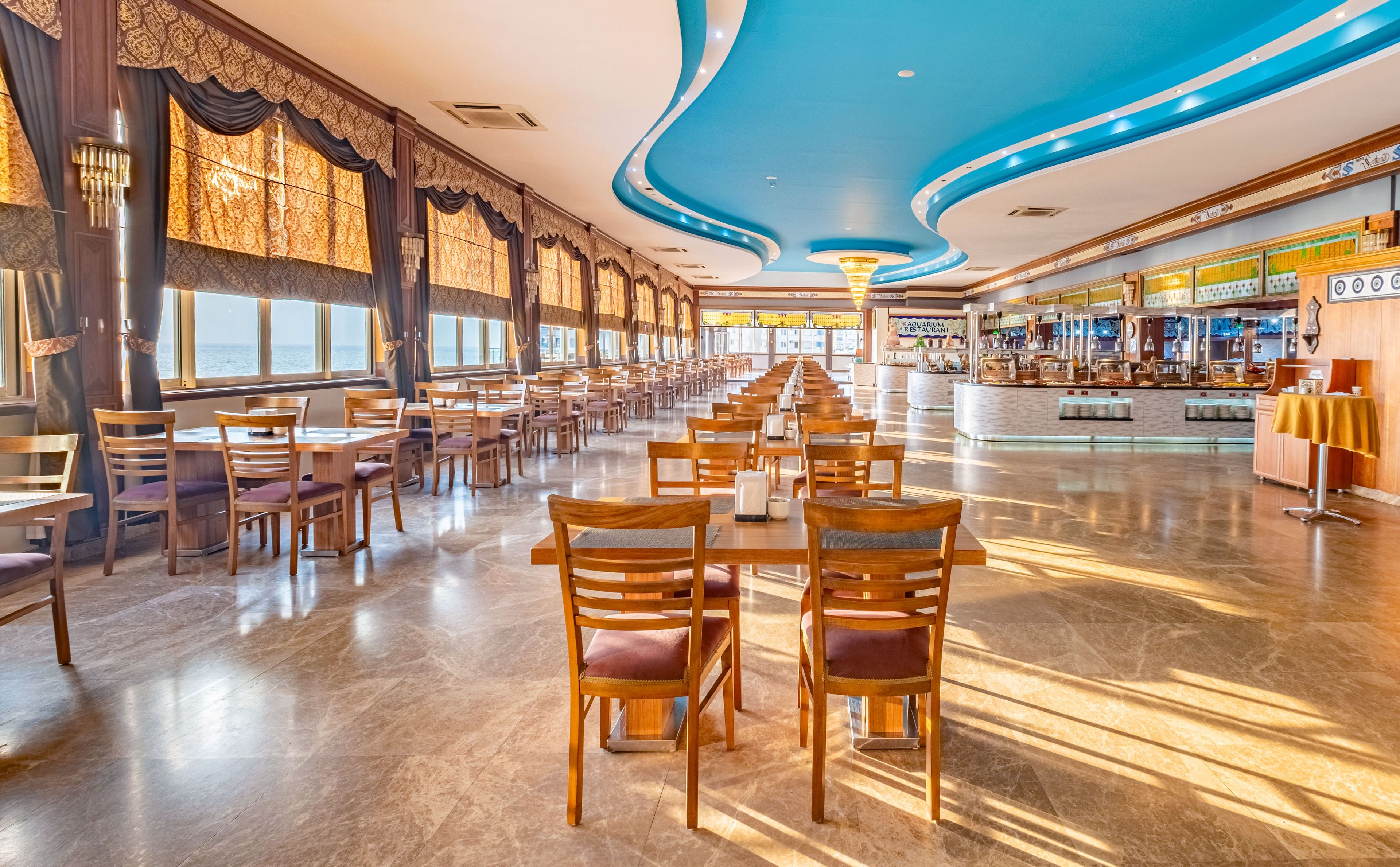 En rymlig restaurang med långa rader av träbord och stolar, stora fönster med utsikt över havet och ett blått tak.