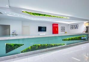 Een moderne receptiebalie met een glazen toonbank, plantenarrangementen met groene planten, een rode deur en een wandgemonteerde televisie.