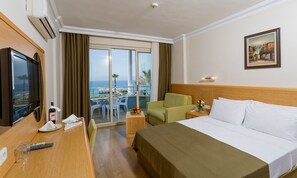 Ein Hotelzimmer mit Bett, Fernseher, Schreibtisch mit Stuhl, einem kleinen Tisch mit Flasche und Gläsern, einem Sessel, einem Balkon mit Meerblick und einem Bild an der Wand.