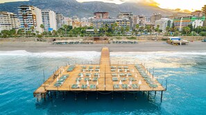 Une jetée en bois, agrémentée de chaises longues et de parasols, s’avance dans la mer, avec un paysage urbain et des montagnes en arrière-plan.