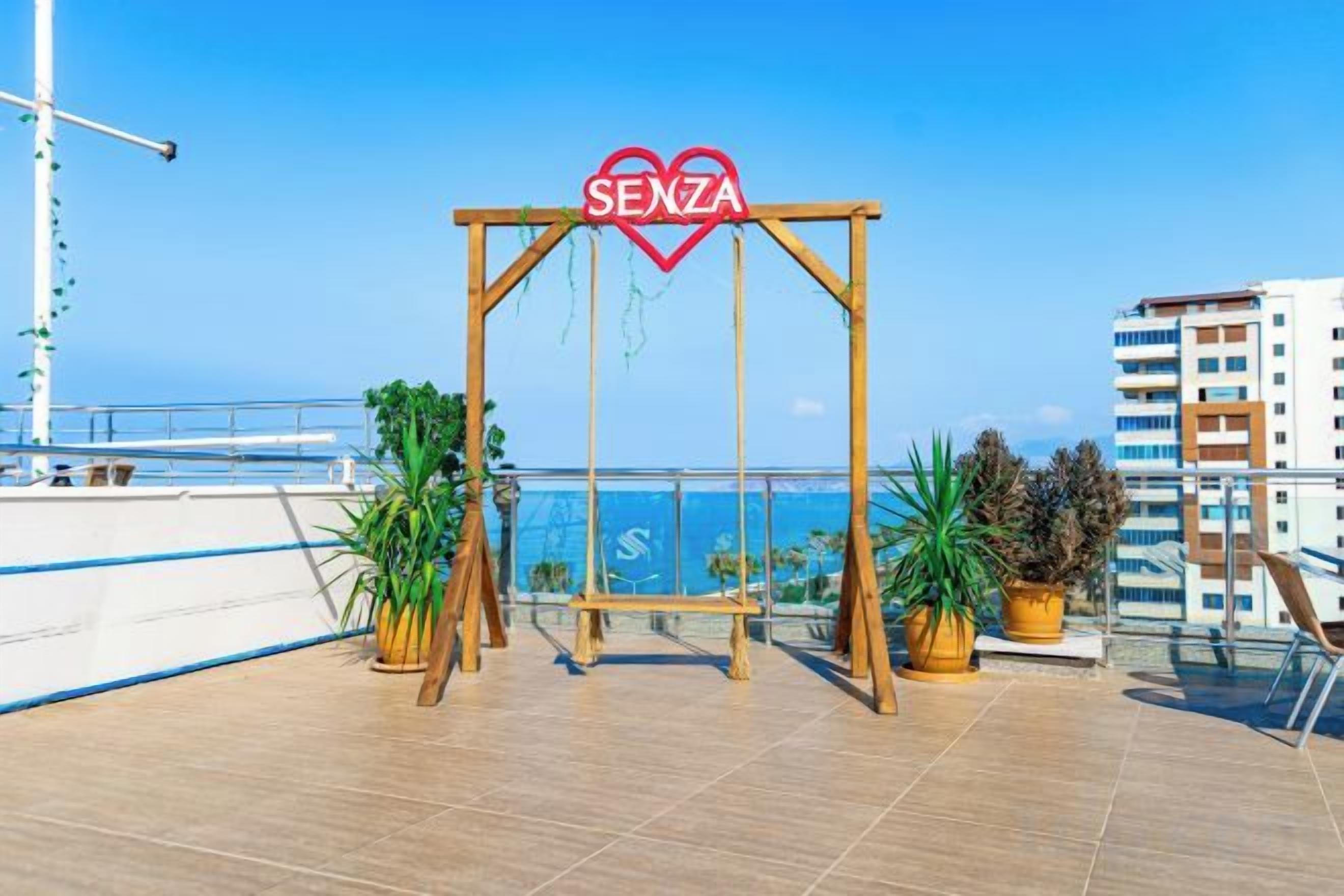 Un columpio de madera con un letrero con forma de corazón que dice 'SENZIA' en una terraza en la azotea con plantas en macetas y vista al mar.