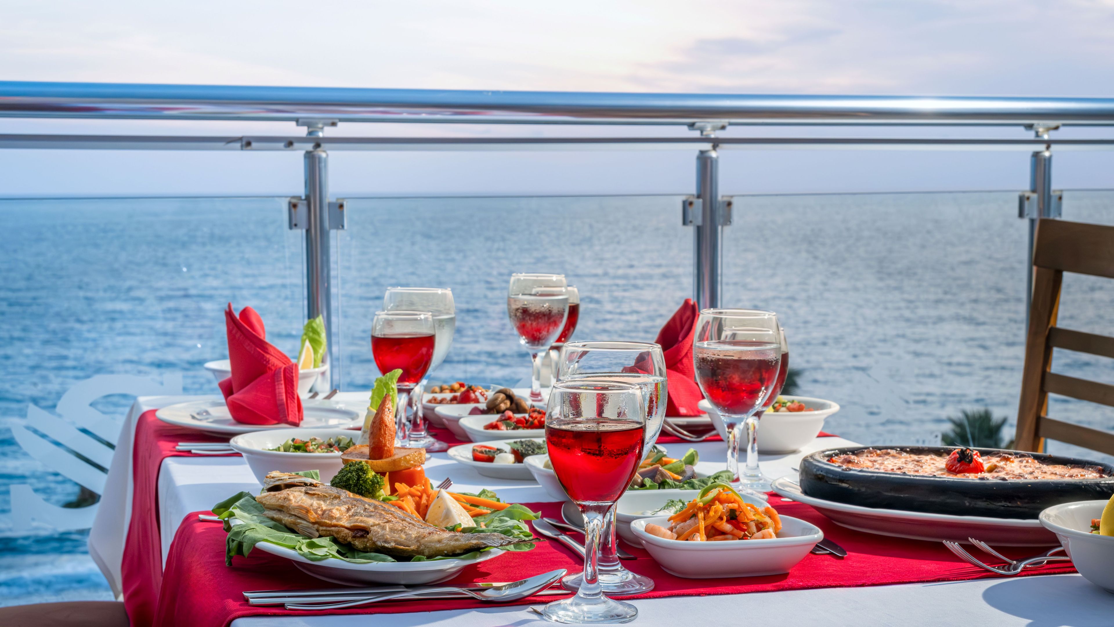 A table set with various dishes, glasses of red wine, and a view of the sea.