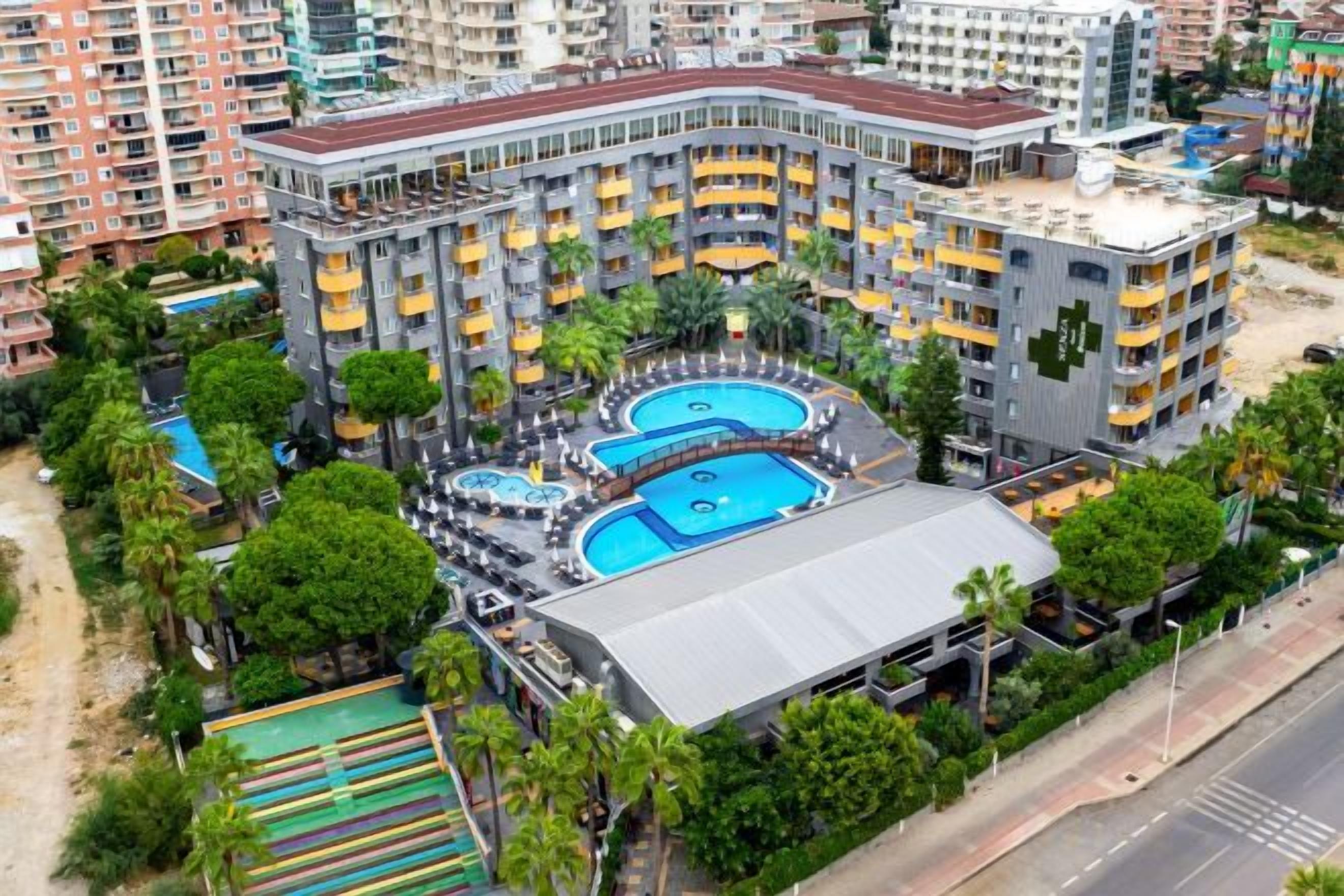 Un hotel de varios pisos con piscina en la azotea, rodeado de vegetación y contiguo a una calle.