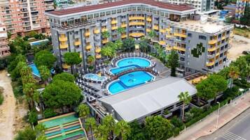 Un hotel de varios pisos con piscina en la azotea, rodeado de vegetación y contiguo a una calle.