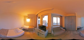 Vue panoramique d’une chambre d’hôtel à l’agencement incurvé, comprenant un lit, un coin salon, un téléviseur et une vue sur la ville à travers de grandes fenêtres.
