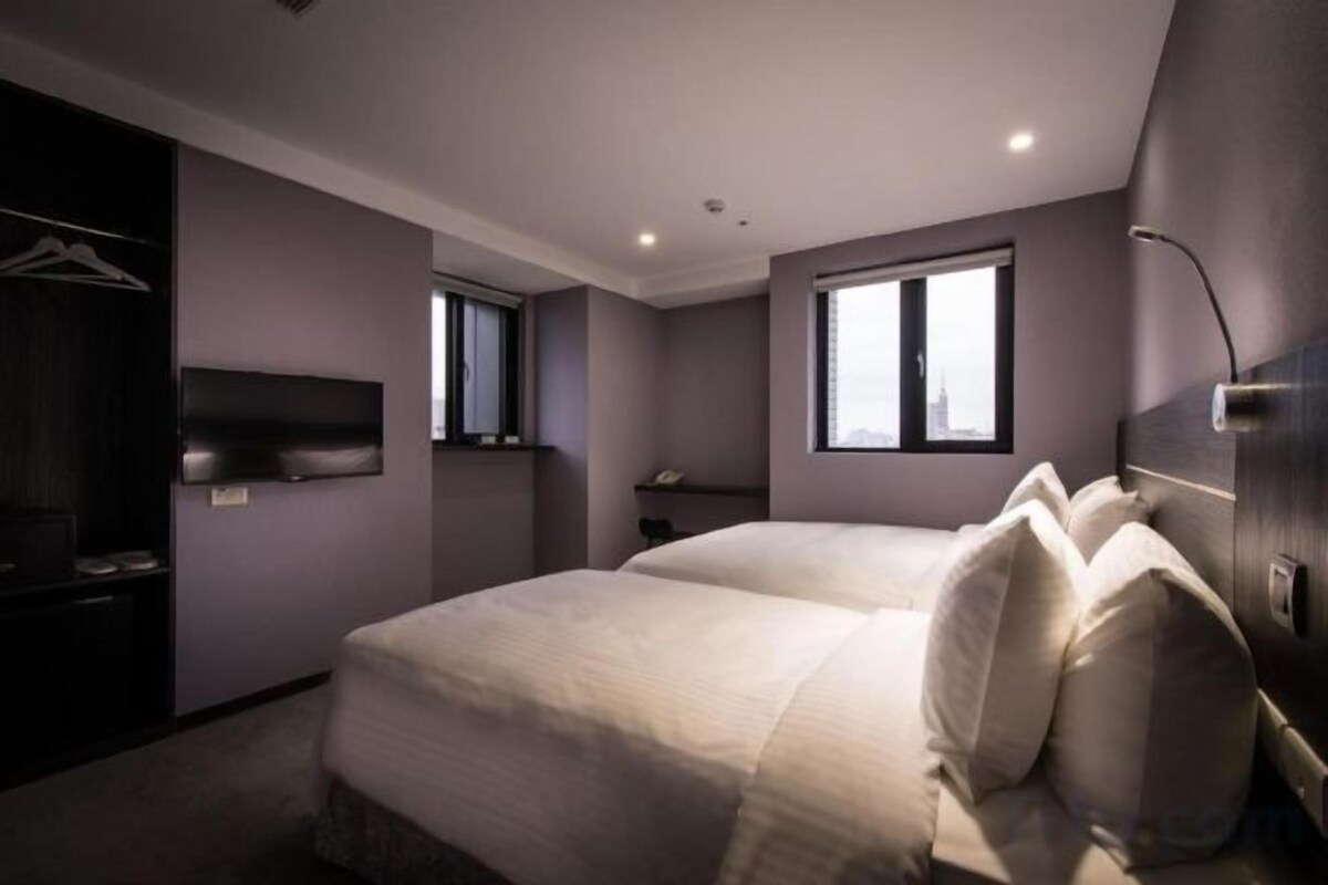A modern hotel room with a large bed, a wardrobe, a flat-screen TV, and two windows.