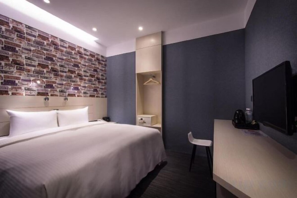 A hotel room with a large bed, a desk, a television, and a brick-patterned wall.