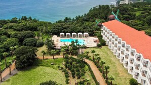 An aerial view of a resort with a swimming pool surrounded by lush greenery, a red-roofed building, and a view of the ocean. - Kensington Resort Jeju Jungmun (Seogwipo)