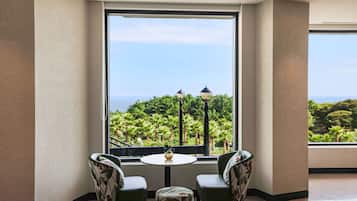 A room with a large window offering a view of a lush green landscape and the sea. There is a round table with two chairs and a small decorative item on it.
