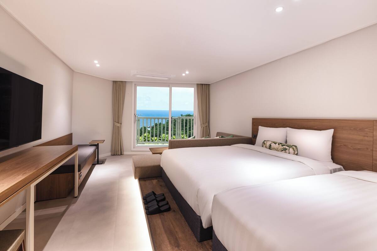 A hotel room with two beds, a TV, a desk, and a balcony with a view.
