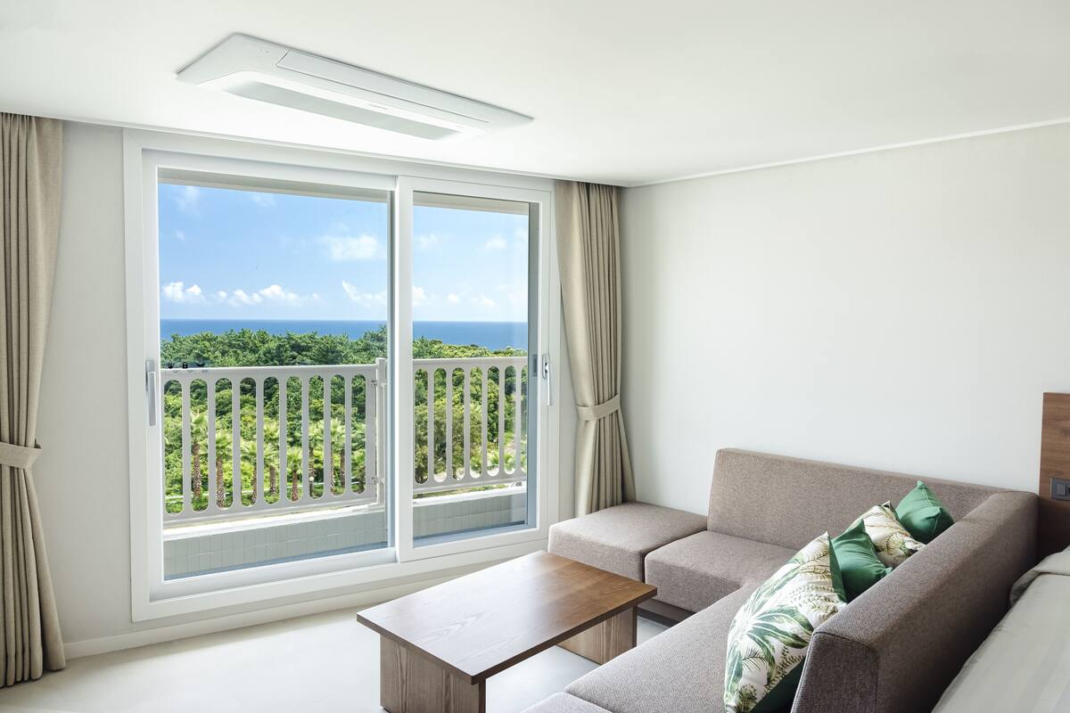 A modern living room with a sofa, a coffee table, and a large sliding glass door leading to a balcony with a view of greenery and the ocean.